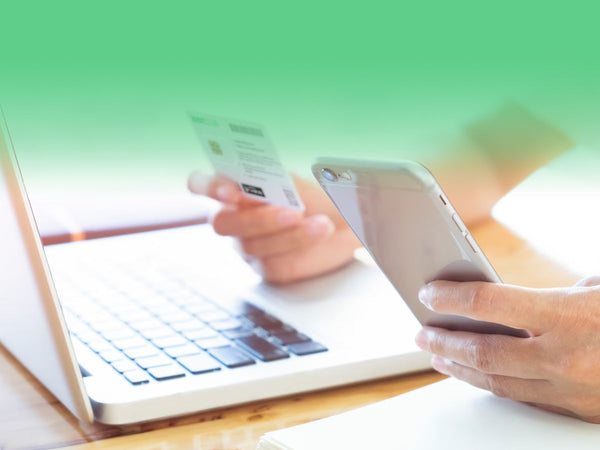 A person holding a smartphone and a card while using a laptop, likely for an online transaction.
