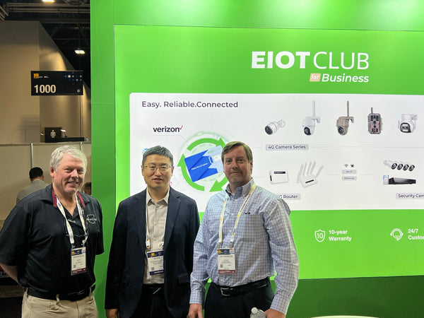 Three men posing for a photo in front of the EIOTCLUB Business booth at a trade show.