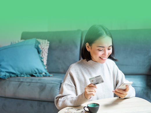 A smiling woman sits in her living room, holding a SIM card and looking at her smartphone.
