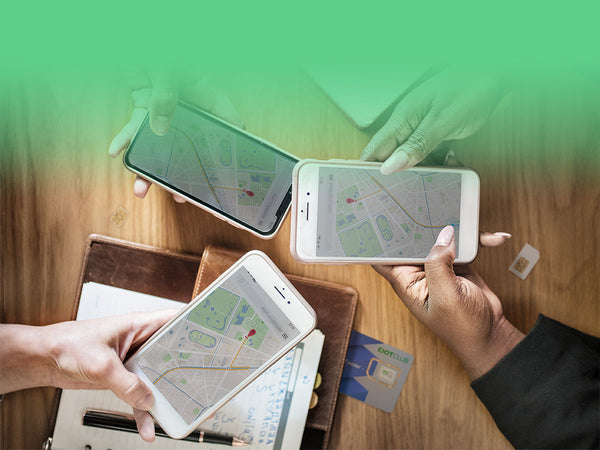 An overhead view of three people holding smartphones, all displaying a map application over a wooden table.