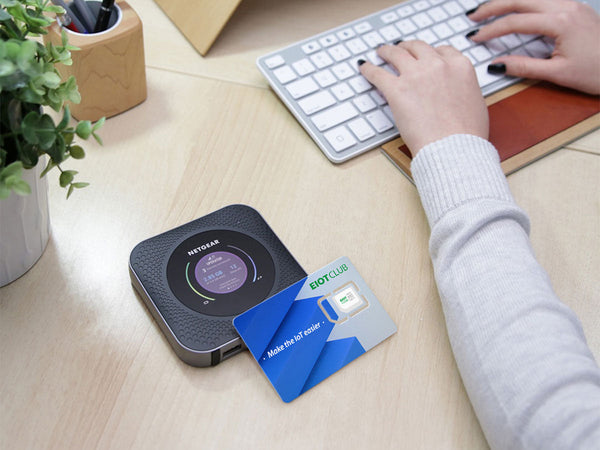 A person typing on a keyboard with a portable Wi-Fi hotspot and an EIOTCLUB SIM card on a wooden desk.