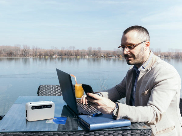 A man working remotely on his laptop and phone at a table by a river, with a portable Wi-Fi router.