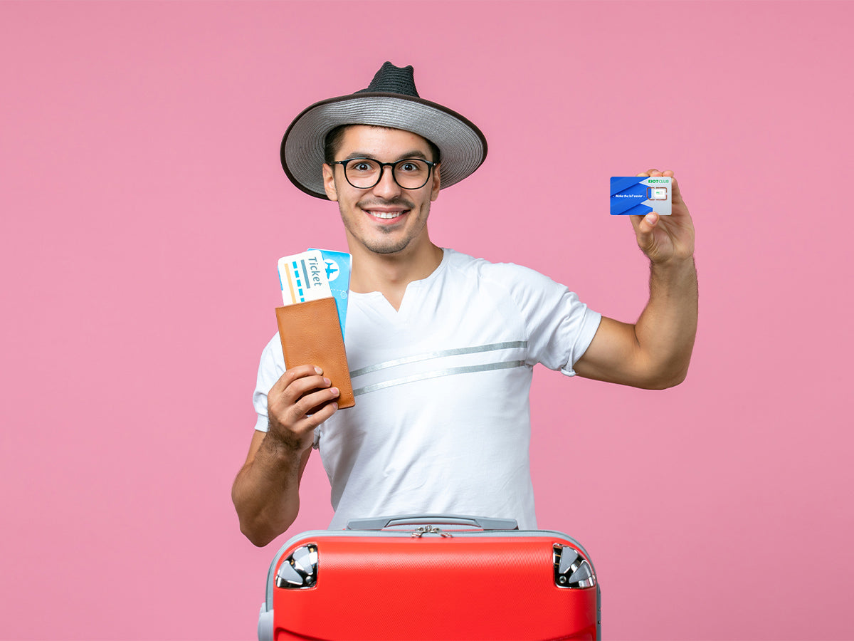 A man in a hat and glasses holds a passport, tickets, and a SIM card while standing with a red suitcase in front of a pink background.