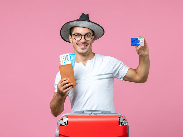A man in a hat and glasses holds a passport, tickets, and a SIM card while standing with a red suitcase in front of a pink background.