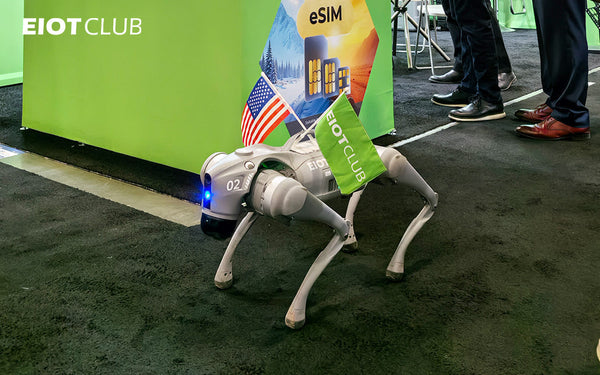 A four-legged robot dog, carrying an EIOTCLUB flag and an American flag, is on display on the carpeted floor of a technology trade show booth.